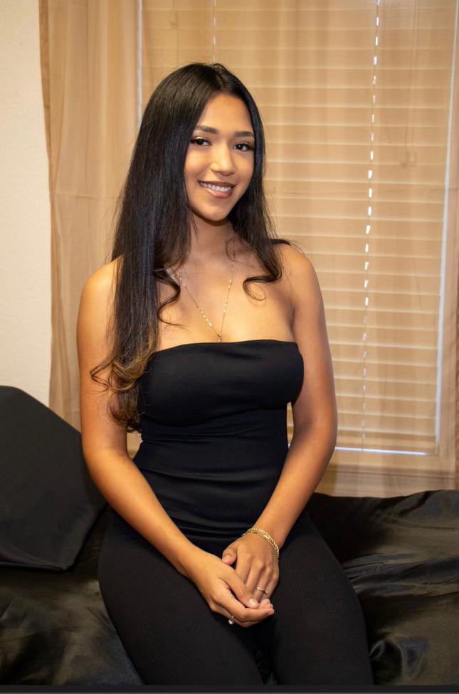 A woman with long dark hair, freshly enhanced by Lakeland Lash and Brow's expert brow tinting, is seated on a bed in a room with beige curtains, wearing a strapless black outfit.