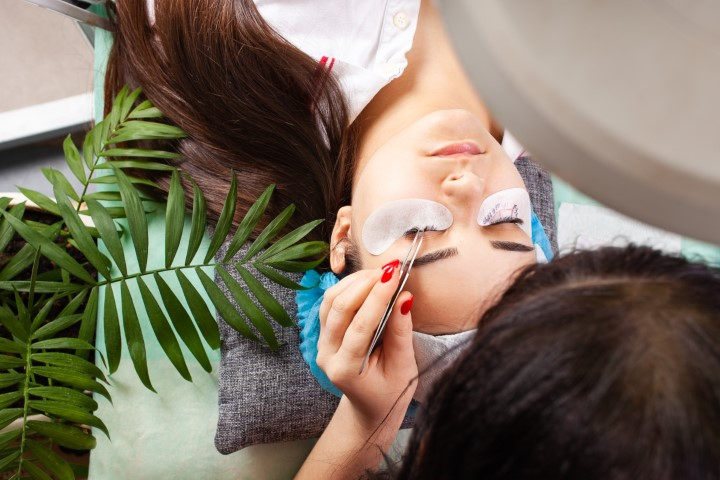 A person is receiving eyelash extensions while lying down, with eye pads placed under their eyes. Another person is applying the extensions using tweezers to help protect your look and maintain lash hygiene.